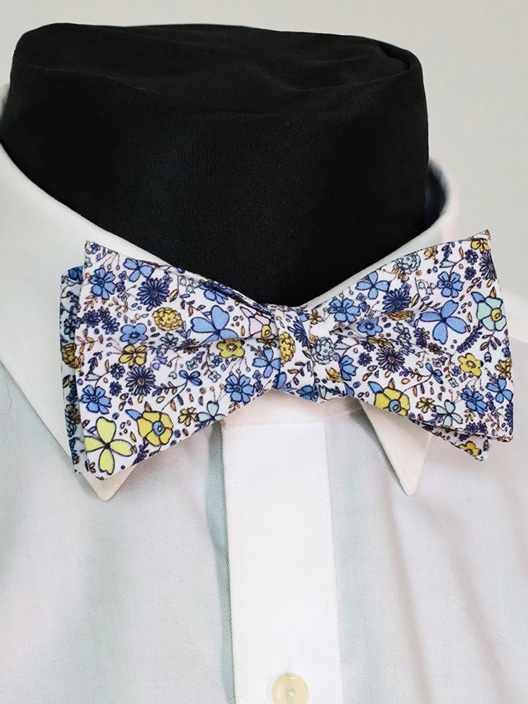 A close - up of a white shirt collar with a multicolored blue floral self - tied bow tie, highlighting its vivid floral pattern.